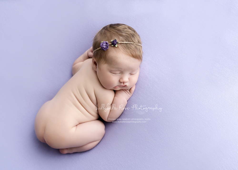 baby sleeping on purple backdrop with rolls and flower headband