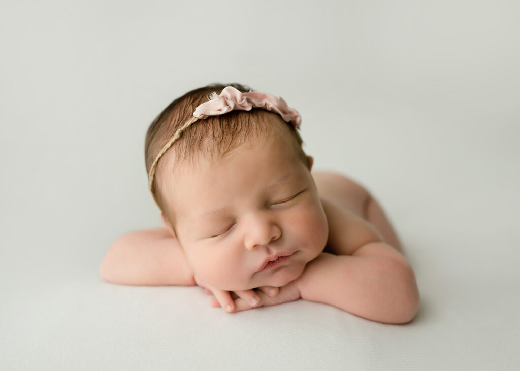 Newborn girl posed on white fabric with her chin on her hands and a pink headband
