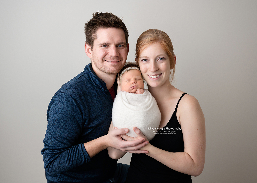 Mom and Dad holding baby girl wrapped in a white swaddle with a pearl headband