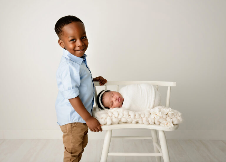 Newborn Photographer, a proud older brother stands beside his newborn baby sister sleeping on a chair.